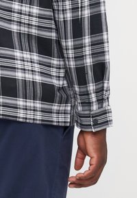 Black and white plaid shirt with long sleeves, featuring a buttoned cuff and a soft, textured fabric. The shirt is tucked into dark blue pants.