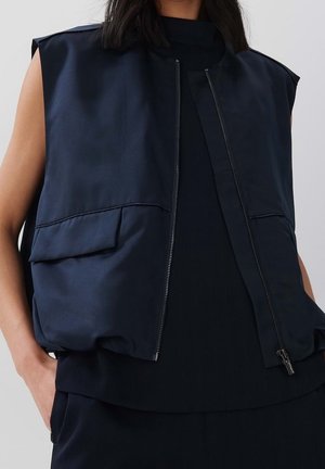 Vrouw draagt een mouwloos marineblauw vest met een rits aan de voorkant en klepzakken, over een marineblauwe top, handen in de zakken, tegen een effen lichte achtergrond.