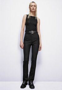 Zwarte mouwloze top gecombineerd met op maat gemaakte zwarte broek. De outfit heeft een slim fit, een glanzende riemgesp en glanzende zwarte schoenen.