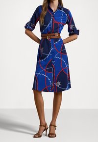 Žena v modré košilové šatech po kolena s červenobílými provázkovými vzory, s dvojitým hnědým p�áskem a hnědými sandály na vysokém podpatku.