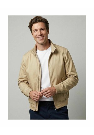 Lachende man met een beige jas over een wit overhemd en een donkere broek, staand tegen een effen lichtgrijze achtergrond.