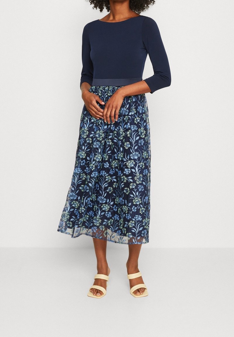 Femme portant un haut bleu marine et une jupe midi fleurie bleu marine avec des sandales beige à bouts ouverts, debout sur un fond blanc uni.