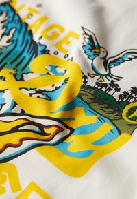 Camiseta de tela blanca con gráficos vibrantes; texto amarillo, olas azules y un diseño de pájaro contra un fondo tropical.