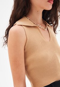 Top senza maniche a coste beige con scollatura a V e colletto. Accentuato da una collana con catena dorata. Texture liscia.