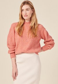 Rosa stickad tröja med texturerade mönster och ribbade manschetter, i kombination med en vit ribbad kjol. Enkel och klassisk design.