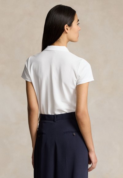 Pikkade tumedate juustega naine, kes kannab valget lühikeste varrukatega polo särki ja tumesinist kõrge vöökohaga pükstesse sisse toppitud, näoga eemale.