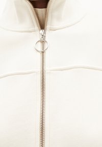 Giacca zip-up crema con una cerniera argentata e un tirante circolare, realizzata in tessuto morbido con una texture liscia e dettagli di cucitura sottile.