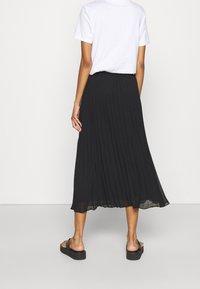Vrouw staat blootsvoets in een zwarte geplooide midi-rok en een witte kortemouwtjes ingestopte blouse tegen een effen achtergrond.