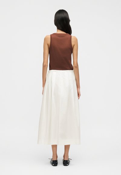 Καφέ αμάνικο τοπ συνδυασμένο με μακριά λευκή φούστα. Η φούστα διαθέτει ελαστική μέση, και το μοντέλο φοράει μαύρα παπούτσια. Μινιμαλιστικός σχεδιασμός.