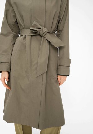Taupefarbener Trenchcoat mit breitem, gebundenem Gürtel, langen Ärmeln mit Manschettenriemen, Händen, die nahe der Seitennaht ruhen, und sichtbaren Taschenkonturen.