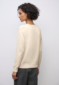 Donna con capelli ricci che indossa un maglione a coste color crema e pantaloni grigi a scacchi, in piedi di lato su uno sfondo chiaro e semplice.