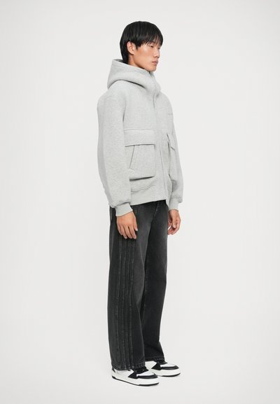 Sweat à zip gris clair avec un col haut et deux poches avant, associé à un jean large noir et des sneakers noir et blanc.