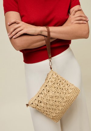 Woman wearing red top and white skirt holds a beige woven handbag with a brown leather strap across crossed arms.