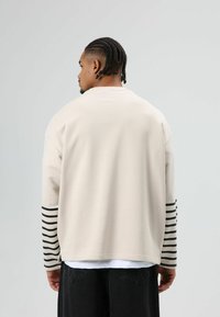 Sweat-shirt beige oversize avec des rayures horizontales noires sur les manches. Confectionné en tissu doux, avec un col rond et une coupe décontractée.