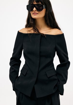 Vrouw met lang donker haar, die oversized zwarte zonnebril draagt en een nauwsluitend zwarte blazer met blote schouders en haar handen in de zakken heeft.