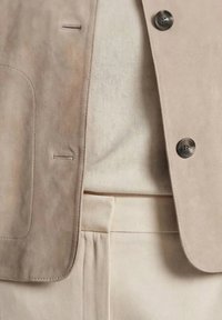 Beige ruskindblazer med sorte knapper, båret over en cremefarvet top, kombineret med cremefarvede bukser med høj talje.