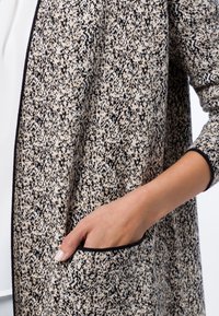 Lekki kardigan z dzianiny w beżowo-czarny wzór. Posiada długie rękawy, otwarty przód oraz boczną kieszeń z czarną lamówką.