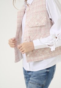 Gilet in tweed rosa con tessuto testurizzato, tasche frontali e chiusura con bottoni, indossato sopra una camicia bianca con polsini legati e jeans azzurri chiari.