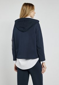 Granatowy krótkotrwały hoodie z kapturem na sznurki i boczną kieszenią. Nosi się go na białej koszuli z zaokrąglonym dołem.