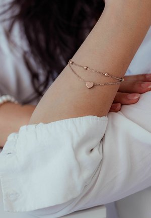 Delicate rose gold double chain bracelet with heart charm and beads worn on the wrist over a white long-sleeve shirt.