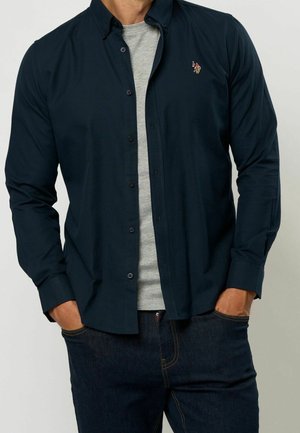 Mężczyzna w ciemnogranatowej koszuli zapinanej na guziki, na jasnoszarej koszulce i ciemnoniebieskich dżinsach, z rękami w kieszeniach, stojący na jednolitym tle.