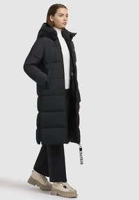 Zwarte pufferjas met horizontaal doorgestikte ontwerpen, capuchon en ritssluiting aan de voorkant. Model draagt beige stevige laarzen en zwarte broek.