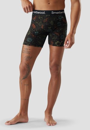 Hombre con bóxers negros con un patrón floral colorido y cintura Smartwool, de pie descalzo contra un fondo liso.