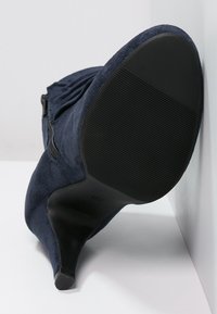 Granatowe zamszowe botki na wysokim obcasie z zaostrzonym noskiem, bocznym zamkiem błyskawicznym oraz teksturowaną czarną gumową podeszwą z poziomymi rowkami zapewniającymi przyczepność.