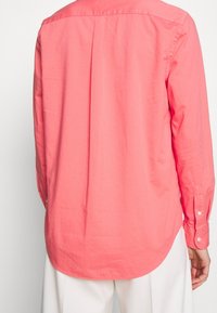 Korallfärgad långärmad skjorta med en plissé bak, knappade ärmslut och rundad nederkant. Tillverkad av slät bomullstyg, med en enfärgad design utan mönster.