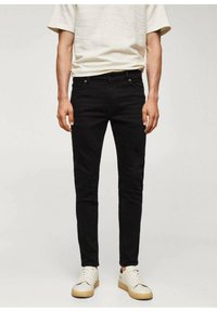 Jean slim noir avec une texture lisse, cinq poches et des éléments en métal, assorti à des baskets crème et une chemise légère et texturée.