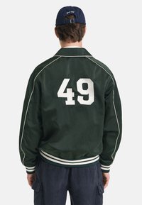 Giacca bomber verde con piping bianco, caratterizzata da un grande numero "49" bianco sul retro, polsini a coste e un cinturino a strisce.