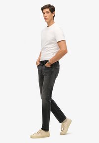 Camiseta blanca de manga corta, jeans negros con una textura ligeramente descolorida y zapatillas beige. Se lleva un reloj en la muñeca izquierda.