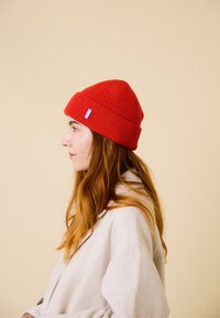 Red knit beanie with ribbed texture and a folded brim. Features a small blue brand label on the side. Simple, close-fitting design.