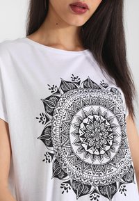 Camiseta blanca con un gran gráfico de mandala negro con patrones intrincados, formas circulares y detalles florales. Textura de tela suave.