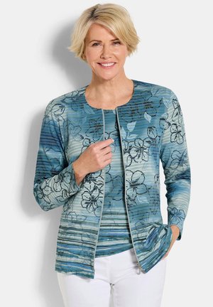 Glimlachende vrouw met kort blond haar, gekleed in een blauwe zip-up jas met bloemenpatroon en een witte broek, staand tegen een witte achtergrond.
