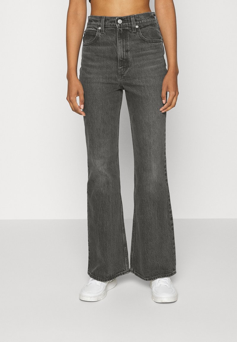 Jeans acampanados de cintura alta en denim gris oscuro con un diseño clásico de cinco bolsillos, combinados con zapatillas blancas. Tejido texturizado con un ligero desgastado.