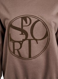 Hnědá mikina s kulatým logem, na kterém je ve tmavě hnědé výšivce nápis "SPORT" a text "Champions keep playing."