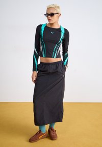 Svart långärmad crop top med turkosa ränder och Puma-logotyp, tillsammans med en svart kjol, turkosa strumpor och bruna sportskor.