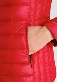 Röd puffjacka med horisontell quiltad design. Har en zippered ficka på vänster sida och en slät, glänsande tygkvalitet.