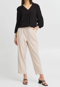 Blouse noire texturée à col en V, à manches longues, associée à un pantalon beige taille haute. Le modèle porte des sandales à talons noires à lanières.