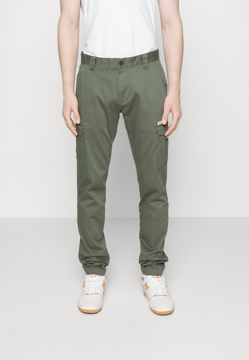Homme portant un pantalon cargo vert olive, un t-shirt blanc et des baskets blanches avec des accents orange, debout devant un fond clair uni.