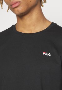 Camiseta de algodón negra con cuello redondo, que presenta un pequeño logo bordado de FILA en rojo y blanco en el lado izquierdo del pecho.