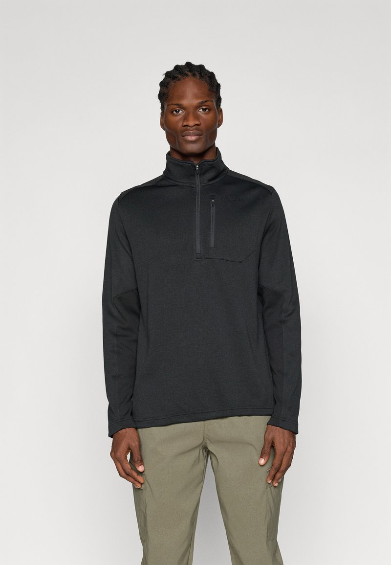 Homme debout face à l'avant portant un pull à fermeture éclair noire et un pantalon vert olive clair sur un fond clair uni.