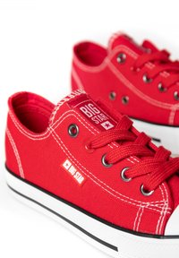 Red canvas sneakers with white rubber soles, featuring black accents, metal eyelets, and a logo tag on the side. Thick red laces.