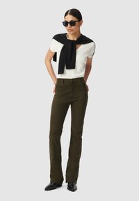 Olive green flared pants with a high waist and button front, paired with a white top featuring embossed text, and a black sweater draped over shoulders.
