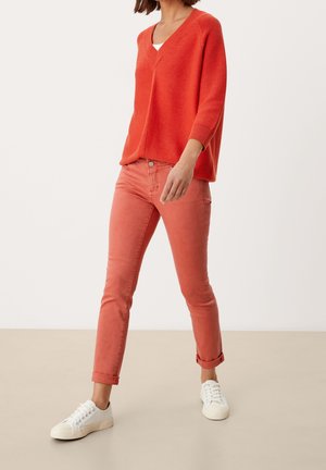 Femme portant un pull en maille rouge ample, un jean slim corail avec les revers roulés et des baskets blanches à lacets, debout devant un fond uni.