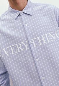 Ljusblå randig skjorta med vita knappar och en framträdande grafisk text "EVERYTHING" i fet stil över framsidan. Tillverkad av bomull.