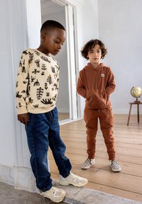 Jongens in casual outfits: de een in een crème trui met zwarte print, donkere jeans en beige sneakers; de ander in een bruine hoodie en joggers met geruite instappers.
