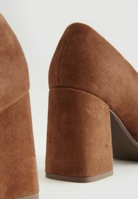 Brown suede high-heeled shoes with a block heel. The texture is soft, and the heel is tapered at the top with a wide, stable base.