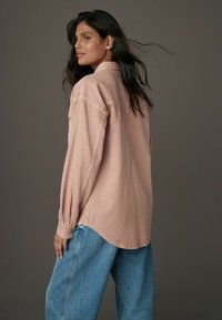 Camicia di jeans a maniche lunghe, rosa chiaro, con colletto e polsini abbottonati, abbinata a jeans blu larghi, su uno sfondo grigio.